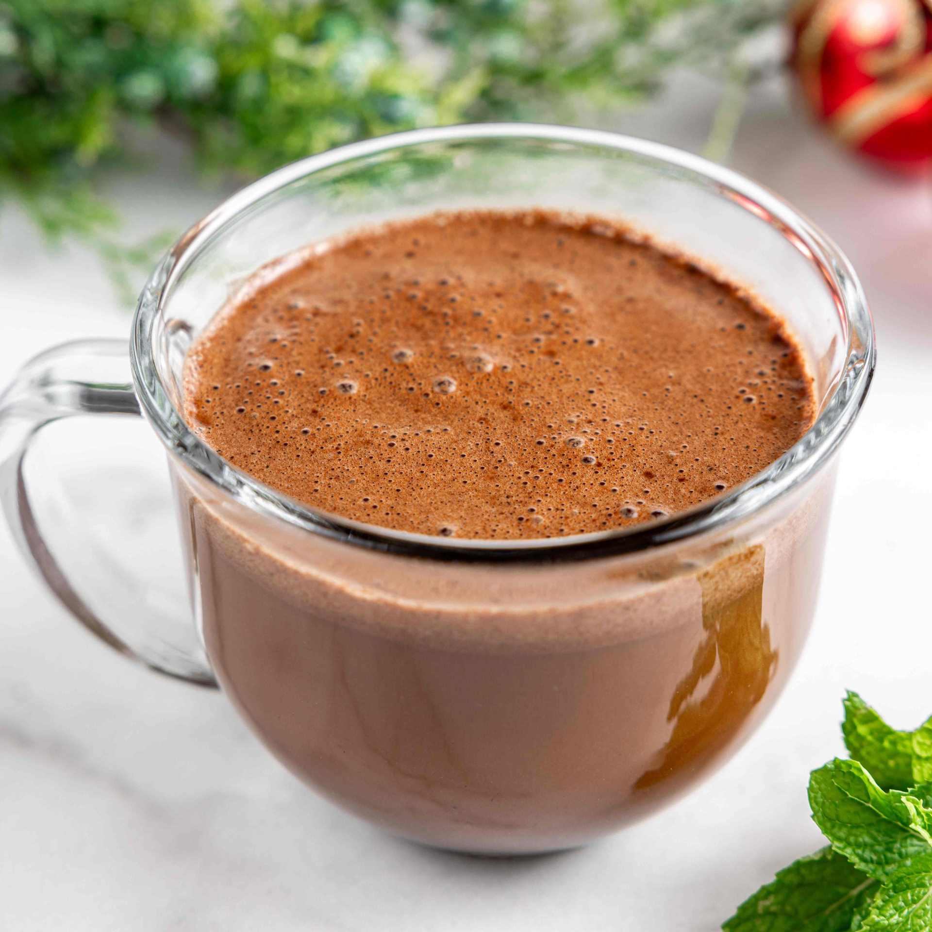 Glass mug of Bare Life vegan peppermint hot cocoa mocha on a marble countertop, garnished with fresh mint leaves and holiday-themed decorations in the background