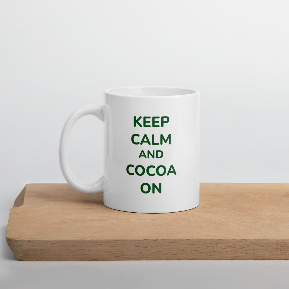 A white ceramic Bare Life mug with the phrase &quot;Keep Calm and Cocoa On&quot; in green lettering sits on a wooden cutting board. The minimalist background emphasizes the sleek design, perfect for cozy hot cocoa moments.