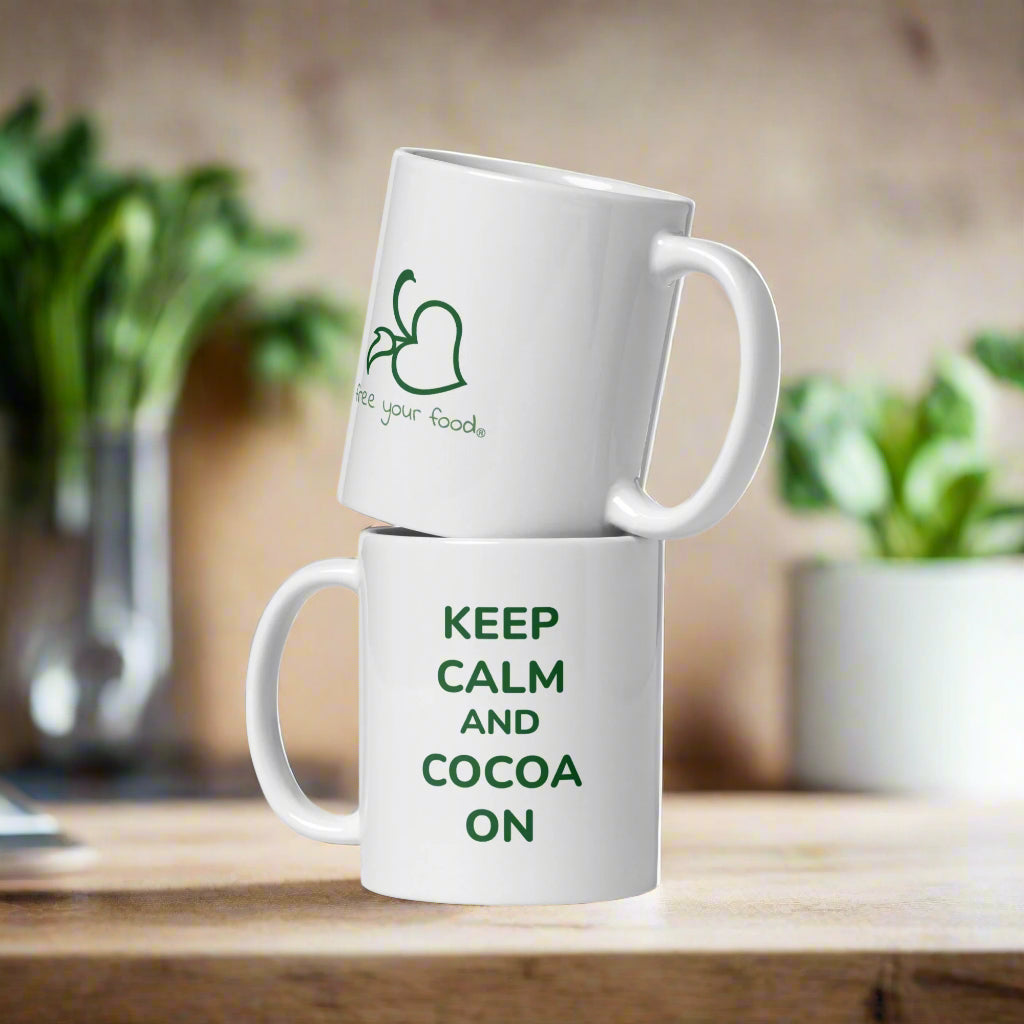 Two white Bare Life mugs stacked on a wooden surface, one featuring the phrase &quot;Keep Calm and Cocoa On&quot; in green text and the other displaying the Bare Life &quot;Free Your Food&quot; logo. A cozy, plant-filled background adds warmth to the scene.