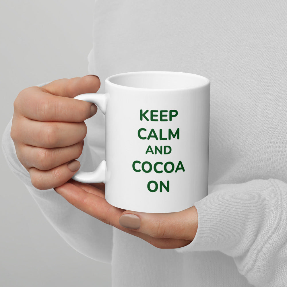 A person wearing a white sweater holds a white Bare Life mug featuring the phrase &quot;Keep Calm and Cocoa On&quot; in green text. The cozy grip and soft background highlight the perfect way to enjoy a warm, dairy-free hot cocoa.