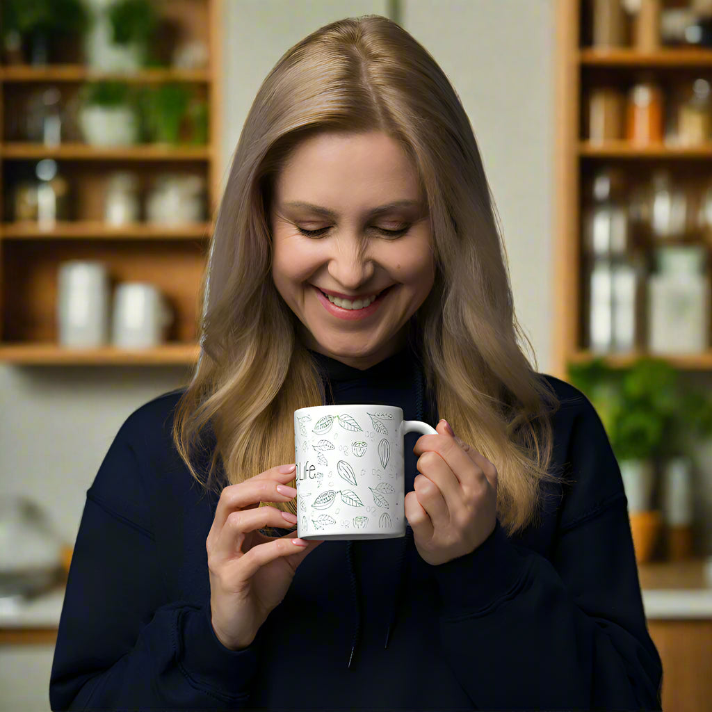 A smiling woman with long blonde hair holds a white Bare Life mug with a botanical design, including leaves and cocoa pods, while standing in a cozy kitchen. She is wearing a dark-colored hoodie and looking down at her drink with a warm expression. The background features wooden shelves with jars, plants, and kitchen essentials, creating a homey atmosphere.