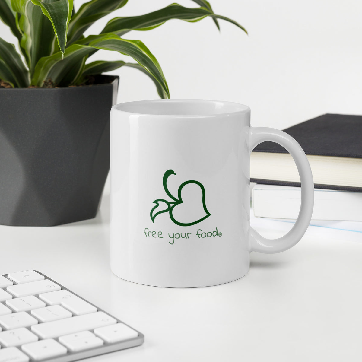 A white Bare Life mug featuring the &quot;Free Your Food&quot; logo sits on a white desk next to a plant, books, and a keyboard. A perfect companion for work breaks, fueling your day with allergen-friendly hot cocoa.